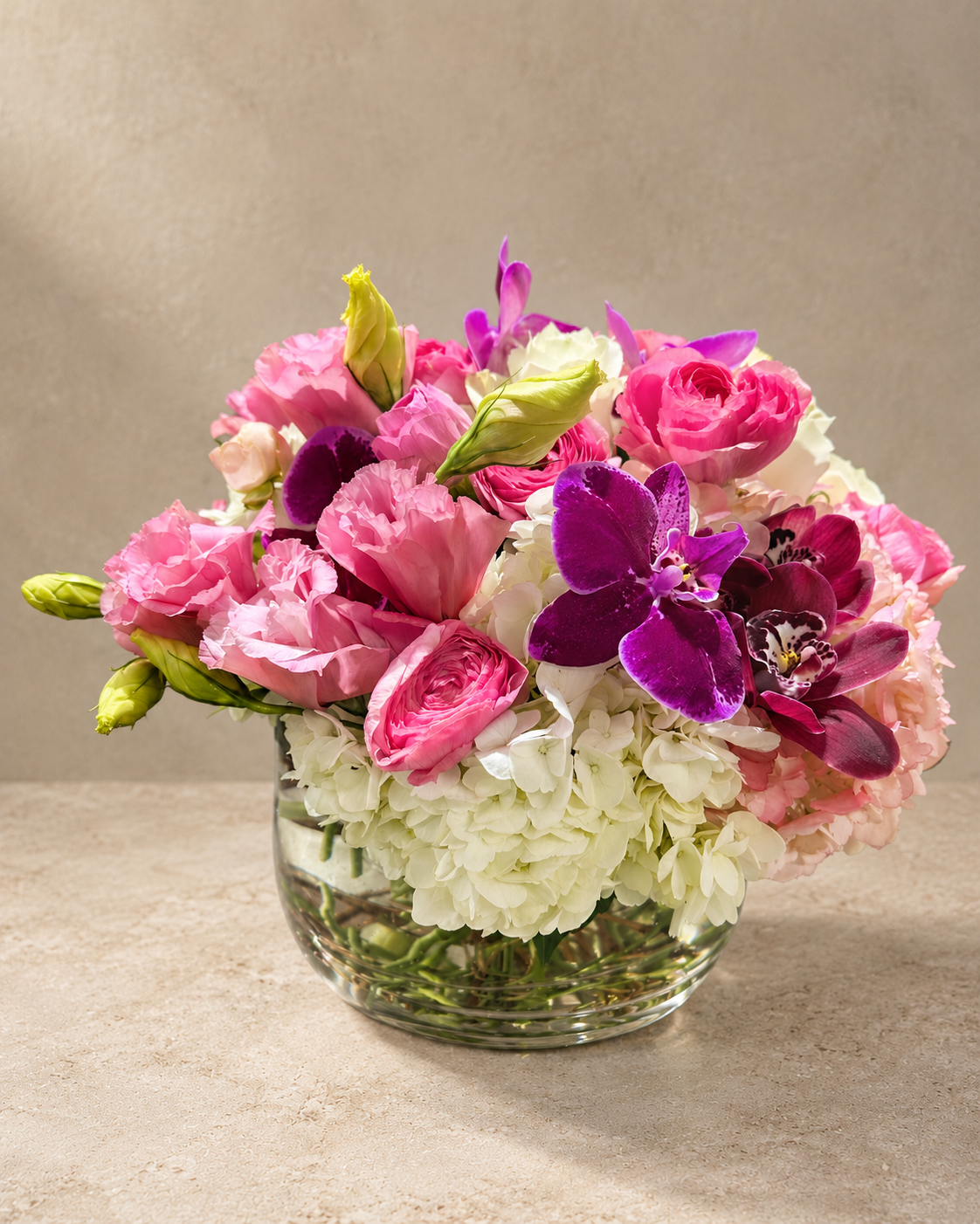 A vibrant floral arrangement featuring pink roses, purple orchids, white hydrangeas, and green buds in a clear glass vase, set against a neutral beige background.