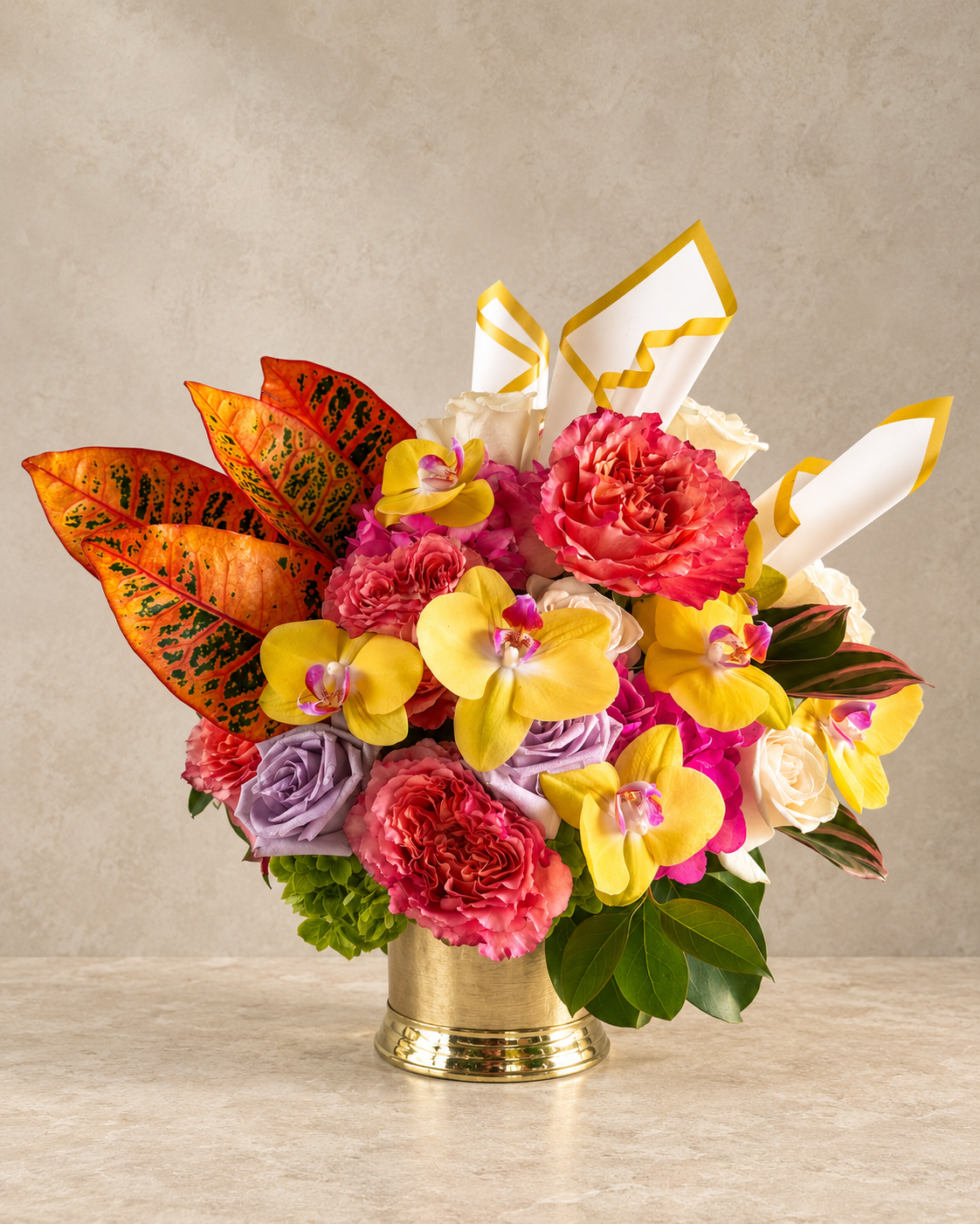 A vibrant floral arrangement in a gold-colored metal vase, featuring yellow orchids, pink and red roses, purple flowers, and orange-green leaves, set against a neutral textured beige background.