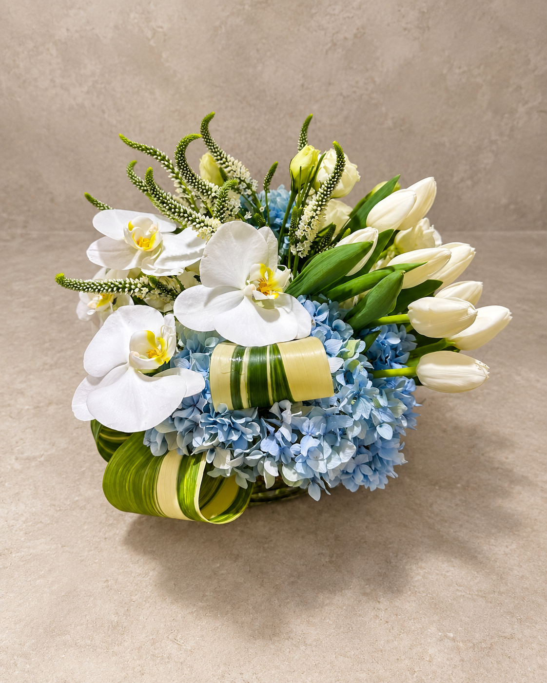 White Butterfly arrangement viewed from above, featuring white orchids, tulips and roses with pale-blue hydrangea, flowering spikes and looped variegated foliage.