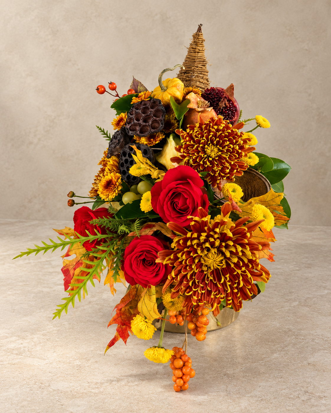 An autumn-themed floral arrangement in a metallic bucket vase, featuring red roses, orange and yellow chrysanthemums, maple leaves, berries, and a small woven cone accent, set against a neutral textured beige background.