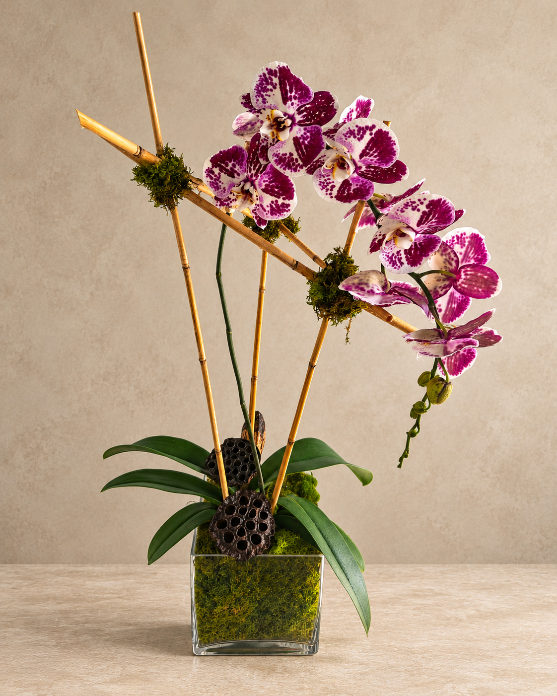 A styled orchid arrangement in a square glass vase with green moss, featuring magenta and white spotted orchids, green leaves, bamboo stakes, and a dried lotus pod against a neutral beige background.