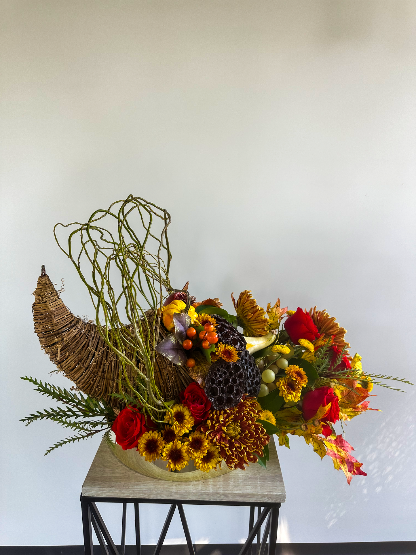 Cornucopia arrangement featuring vibrant red roses, yellow chrysanthemums, and natural elements like lotus pods and mini pumpkins in a festive fall display.