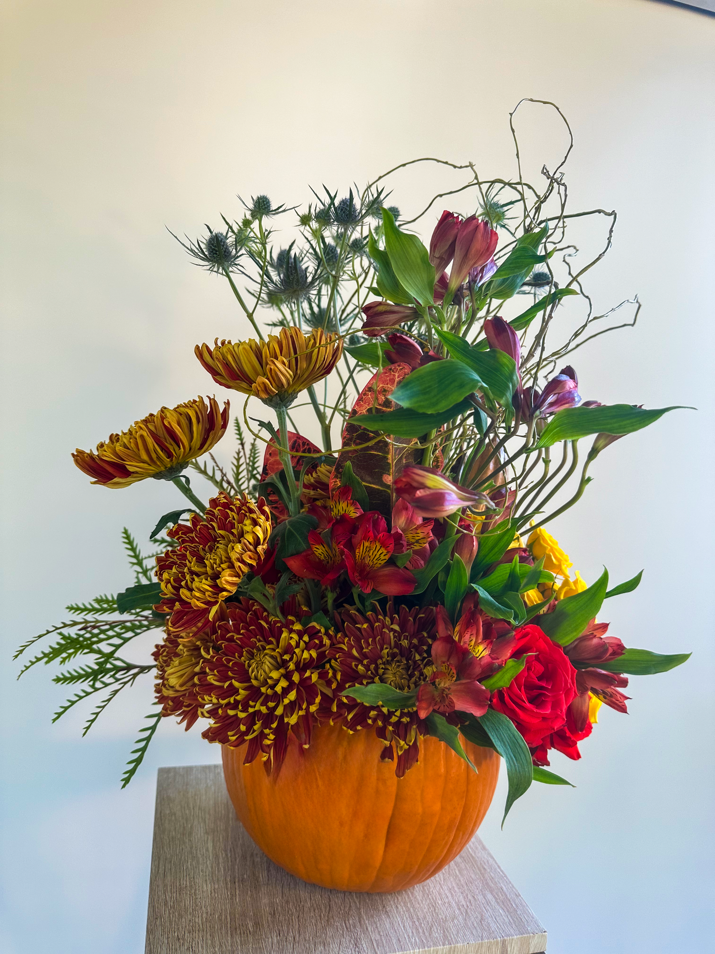 Elegant fall floral display in a pumpkin base, showcasing rich reds, warm yellows, and spiky blue thistle. Designed for seasonal warmth and texture by VIP Floristry, Houston’s luxury florist.