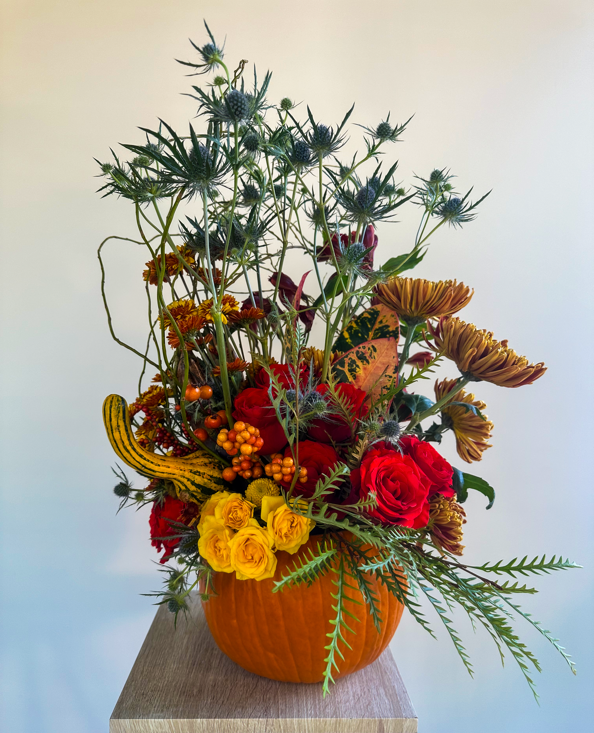Festive autumn floral arrangement in a real pumpkin base, featuring yellow and red roses, vibrant chrysanthemums, and unique dried thistle. A perfect seasonal centerpiece, adding rustic elegance to fall décor by VIP Floristry in Houston.