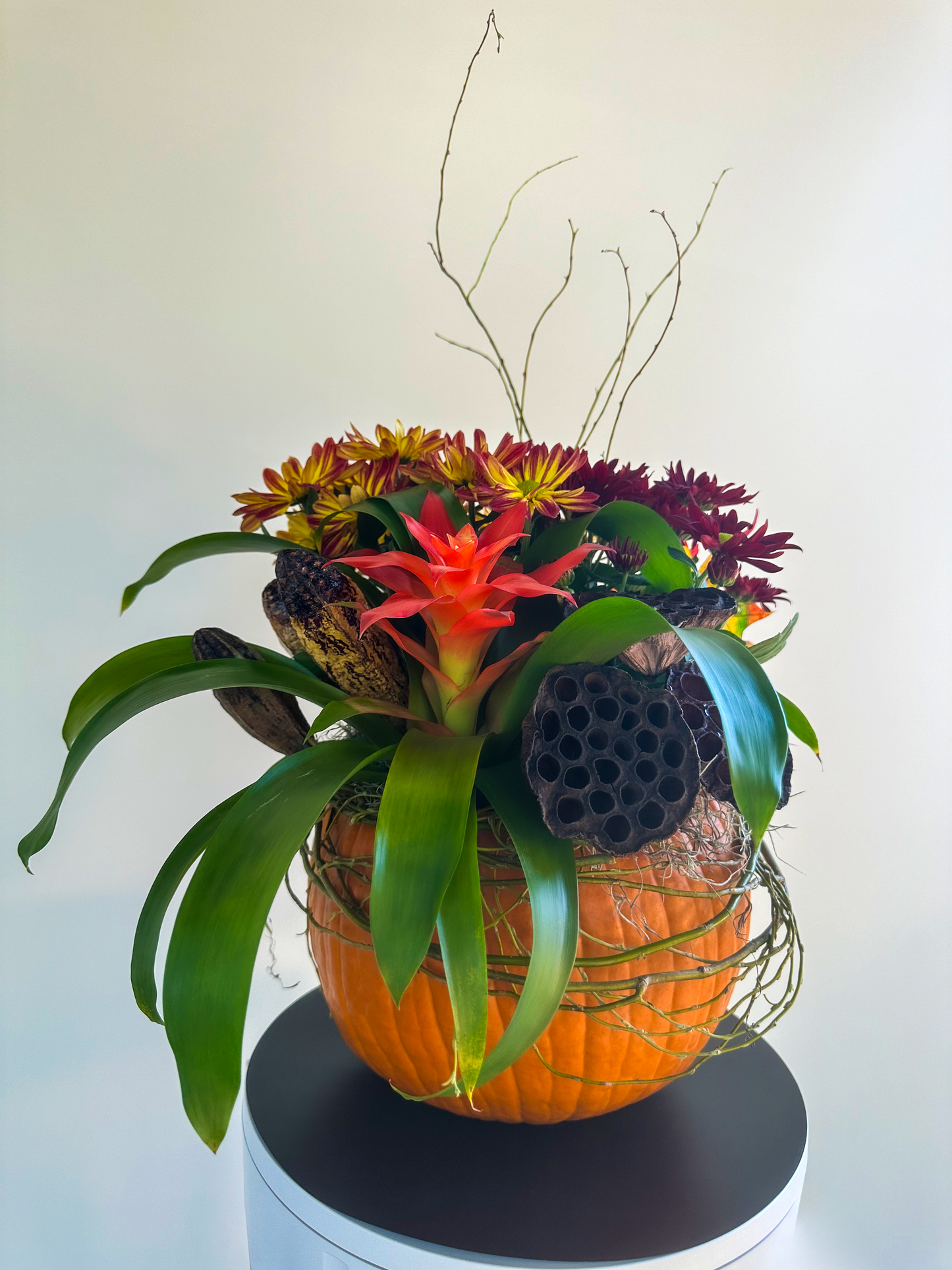 Arrangement in a carved pumpkin featuring a vibrant bromeliad with red flowers, surrounded by golden chrysanthemums and lotus pods, accented with natural twigs. Ideal for autumnal décor or Thanksgiving celebrations.