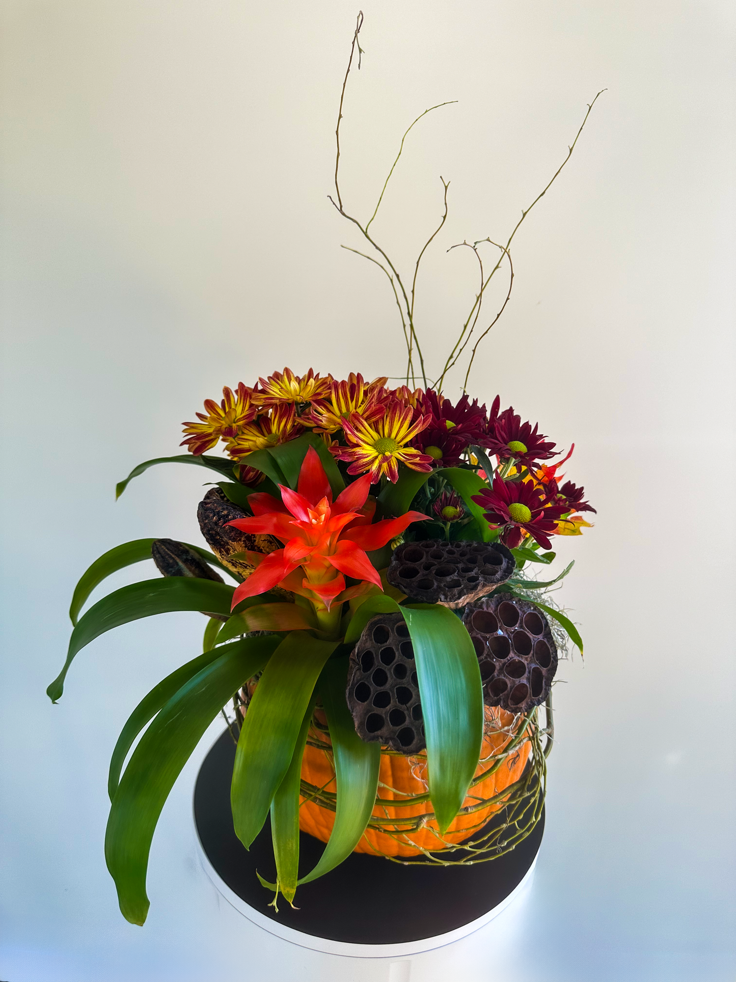 Unique pumpkin-based floral arrangement with a focal bromeliad bloom, lotus pods, and deep red chrysanthemums, accented with curling twigs for height and texture. Perfect seasonal centerpiece for fall.