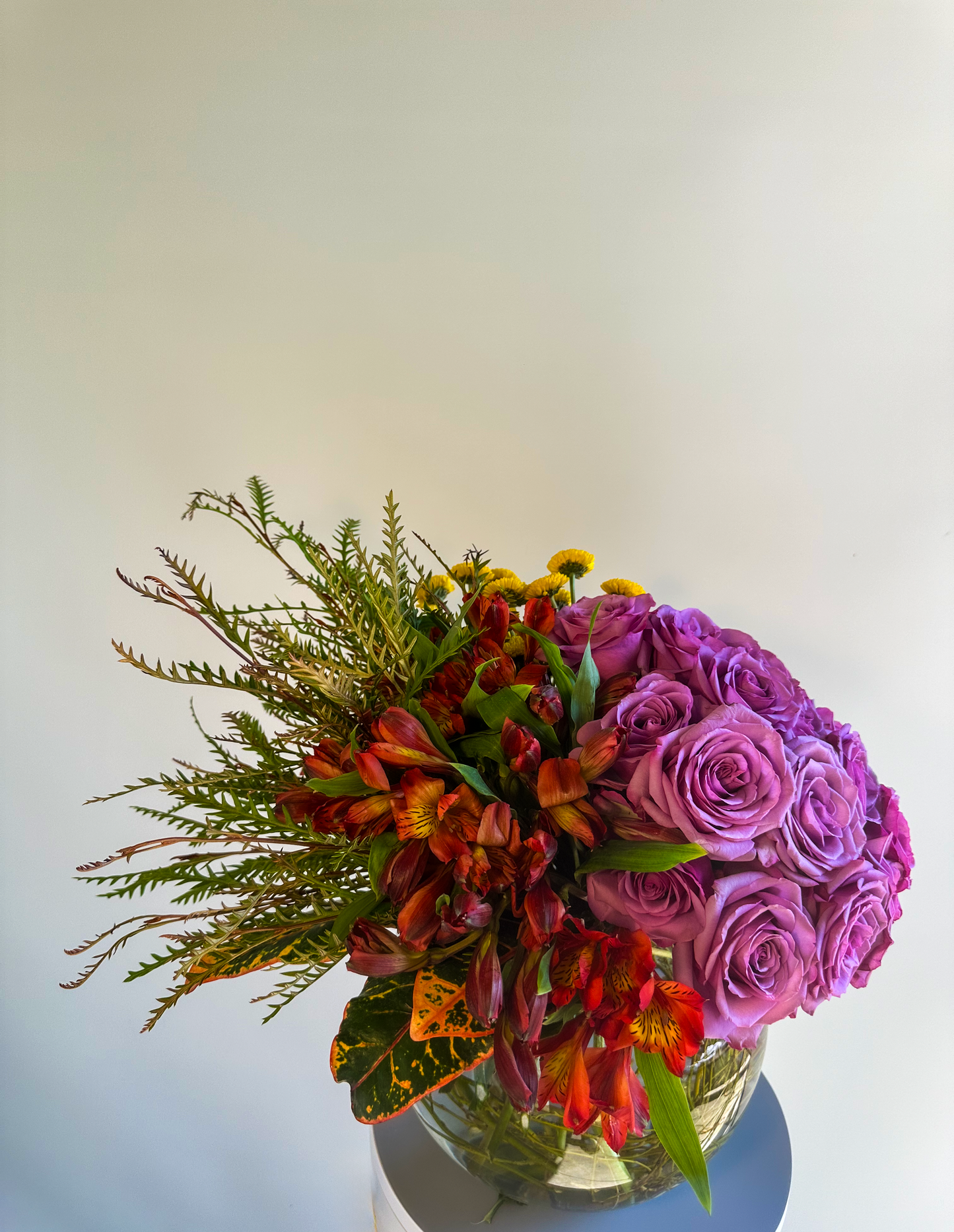 This exquisite floral display includes deep lavender roses paired with rich red alstroemerias and yellow chrysanthemums in a clear glass bowl. The greenery adds texture, creating a dynamic, layered look perfect for luxurious Houston events.