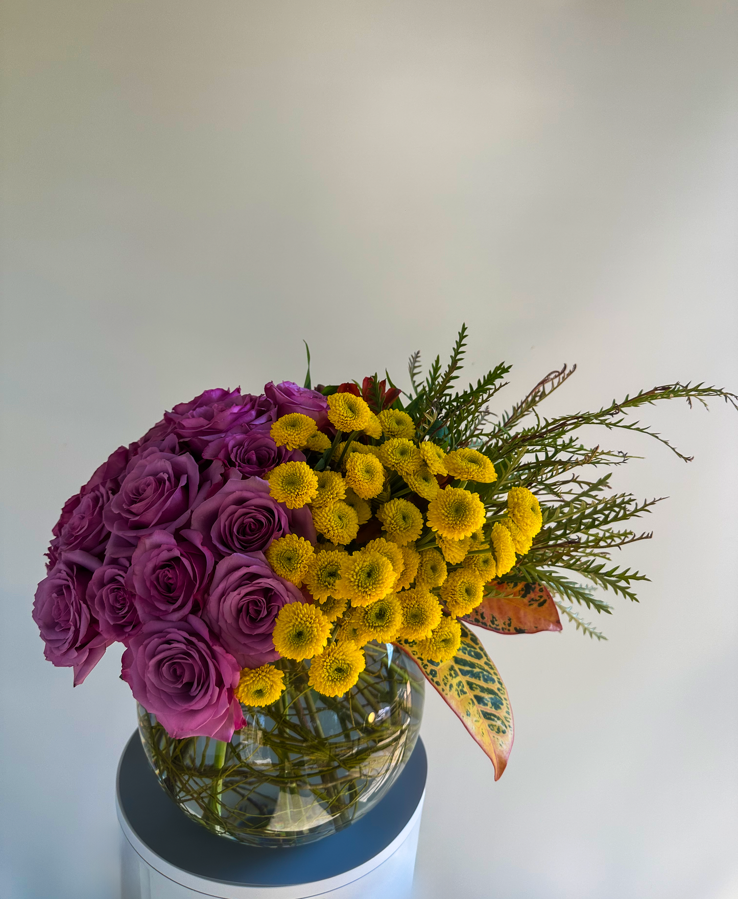 A rounded floral design with vibrant lavender roses and bright yellow button chrysanthemums complemented by tropical leaves and sprigs of delicate greenery in a glass vase. This arrangement, perfect for upscale Houston occasions, blends soft and vibrant hues for a fresh and eye-catching centerpiece.