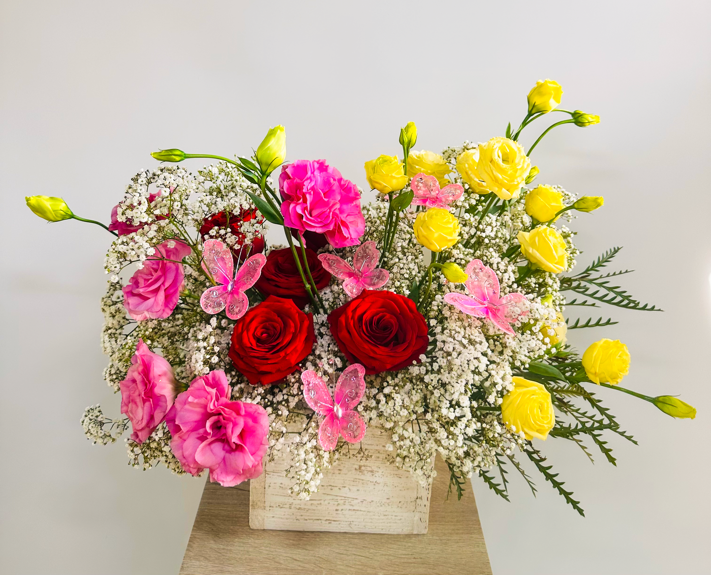 Whimsical floral display with red roses, pink lisianthus, yellow ranunculus, and abundant baby's breath in a rustic wooden vase. Enhanced by delicate pink butterfly decorations for a touch of fairy-tale elegance.