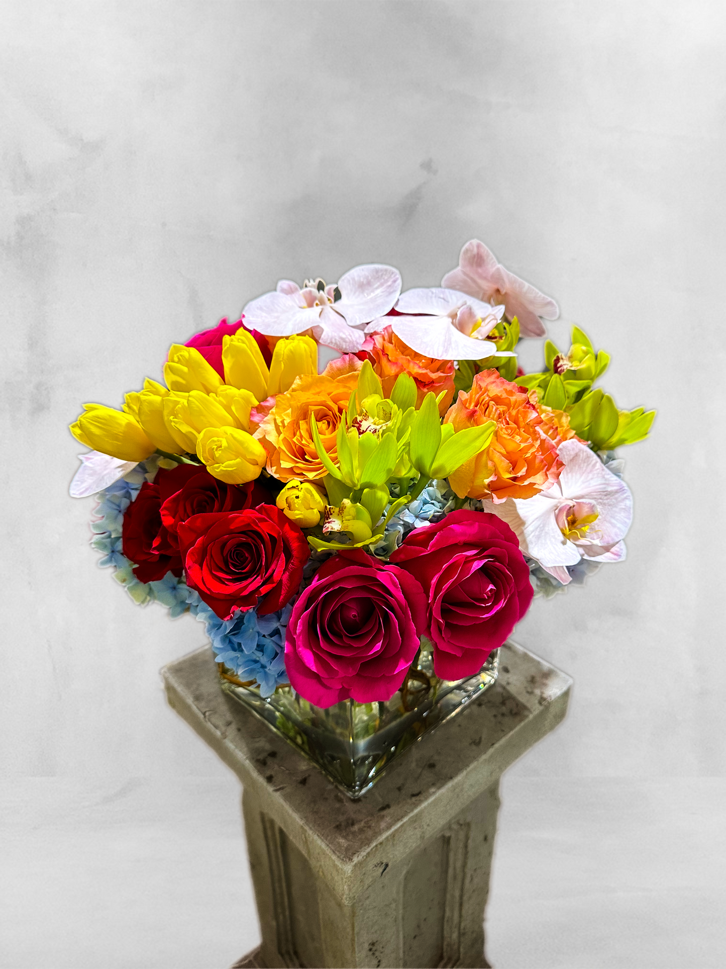 Vibrant floral arrangement with pink and red roses, yellow tulips, orange orchids, and blue hydrangeas in a glass vase, showcasing VIP Floristry's luxury custom designs in Houston.