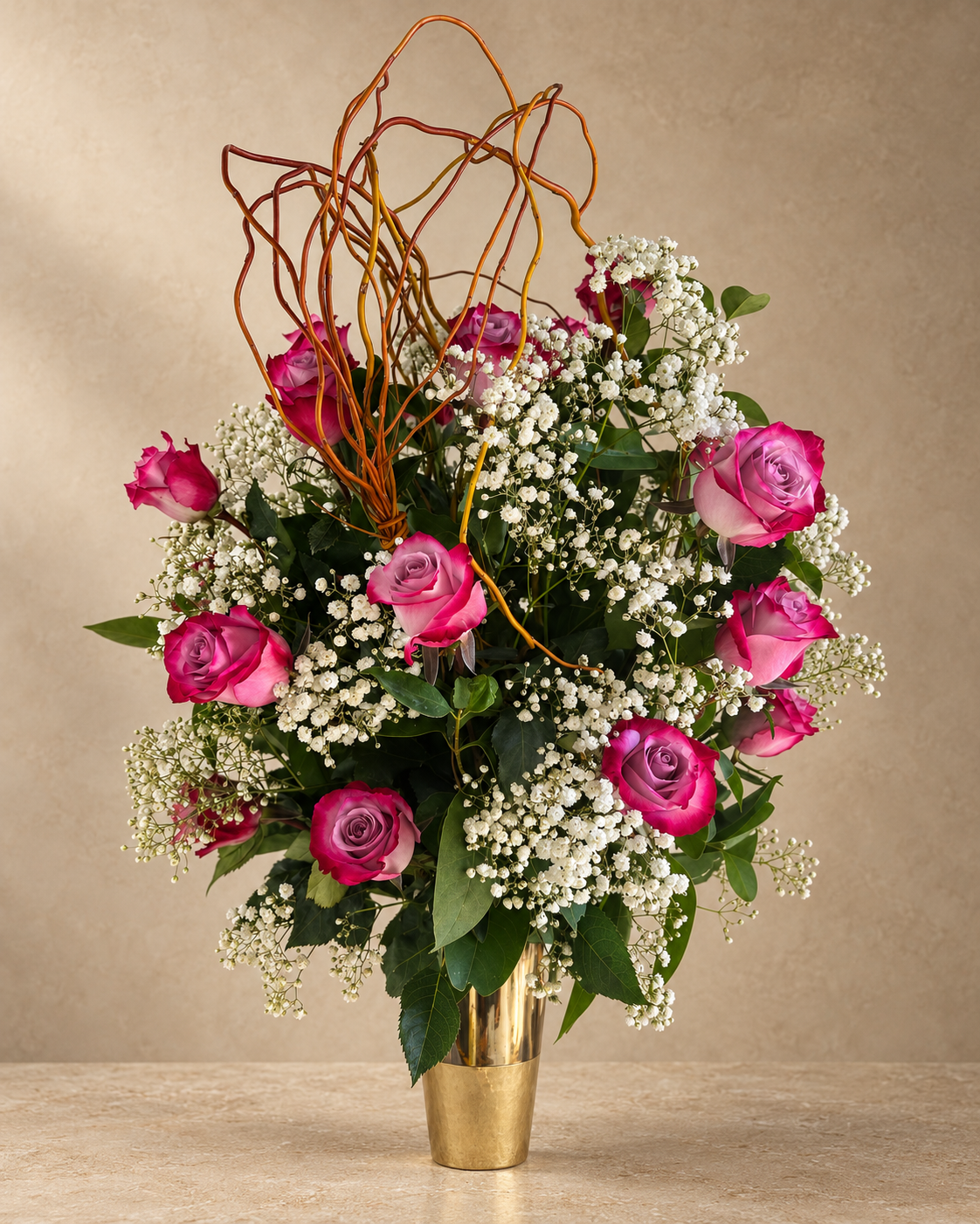 A floral arrangement featuring vibrant magenta-pink roses, delicate white baby's breath, and green foliage, styled in a shiny gold cylindrical vase against a neutral beige background.