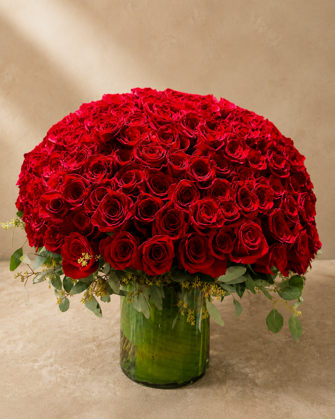 Closer view of a large red rose arrangement with eucalyptus and seeded greenery in a tall green glass vase.