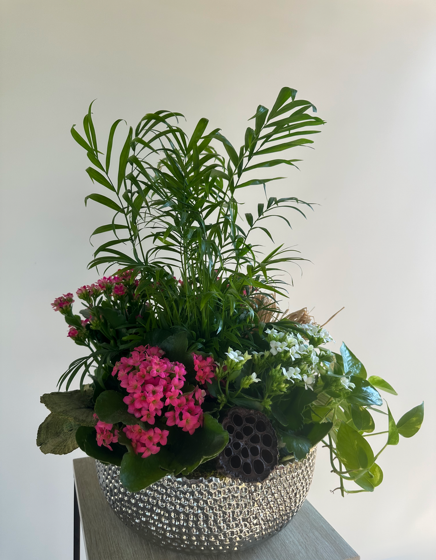 Stylish plant arrangement in a silver textured container with green foliage, pink and white kalanchoe flowers, and lotus pods, designed for luxurious indoor decor in Houston.