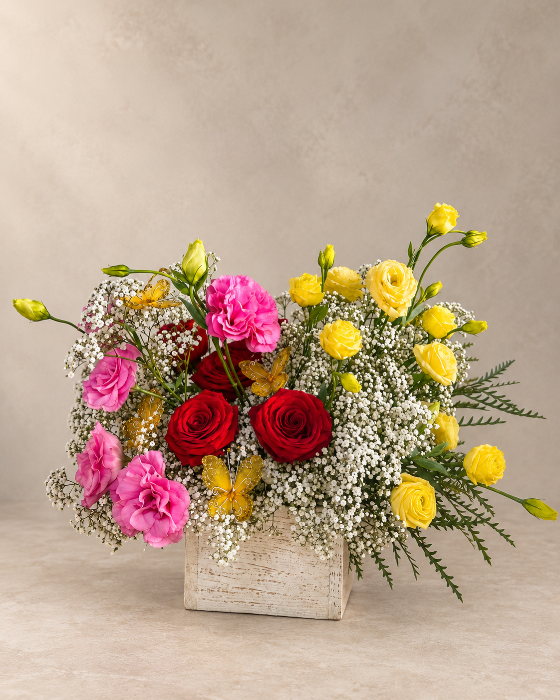 A vibrant floral arrangement in a rustic white wooden box, featuring red and pink roses, yellow roses, pink carnations, and white baby's breath, set against a neutral beige background.