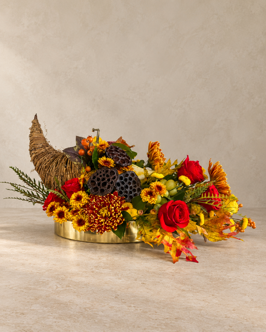 An autumn-themed floral arrangement in a metallic bucket vase, featuring red roses, orange and yellow chrysanthemums, maple leaves, berries, and a small woven cone accent, set against a neutral textured beige background.