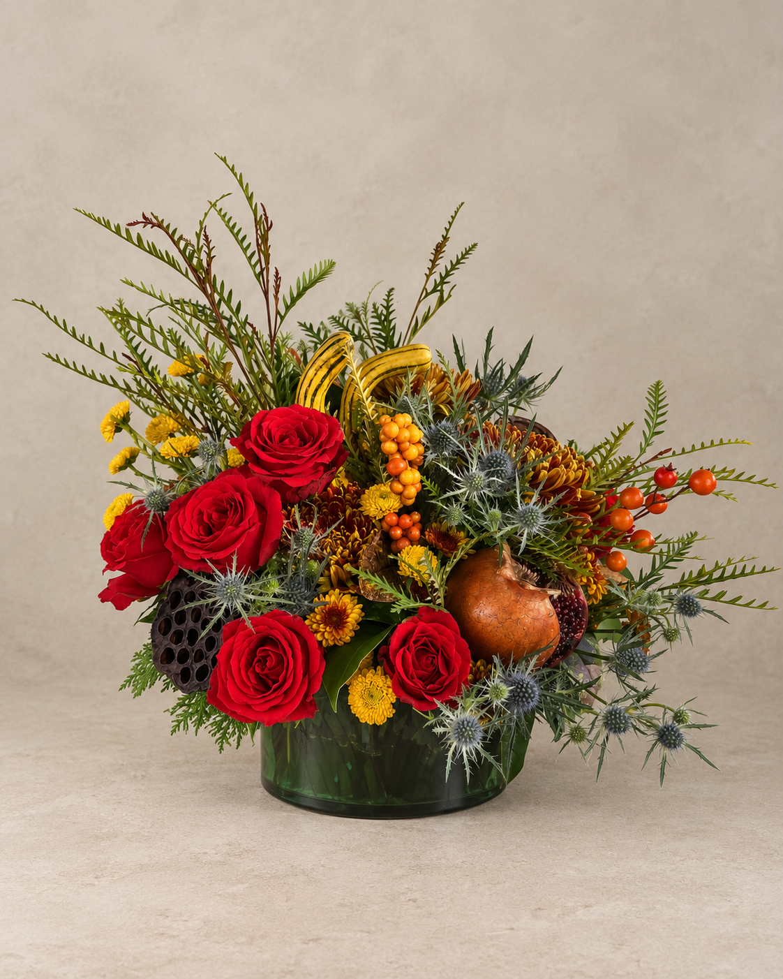 Autumn arrangement with five red roses, bronze chrysanthemums, yellow flowers, blue-gray thistle, striped gourds, berries and a lotus pod in a dark-green glass vessel.