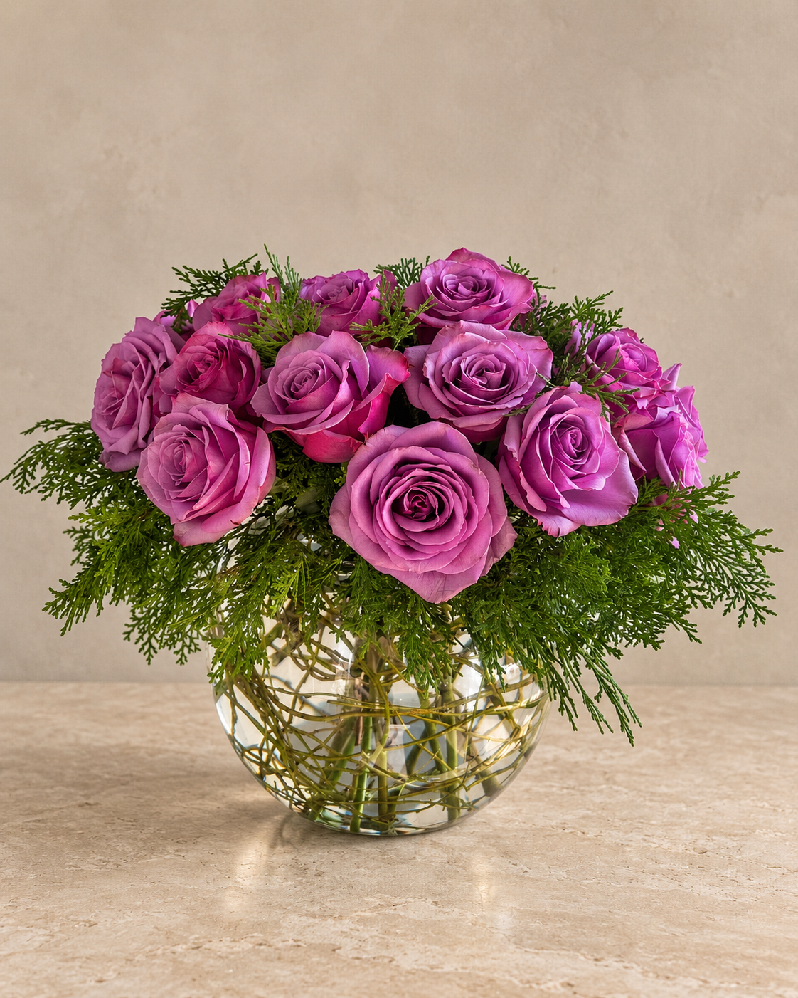 A bouquet of vibrant purple and magenta roses arranged with green foliage in a clear glass vase with a gold wire pattern, set against a neutral beige background.