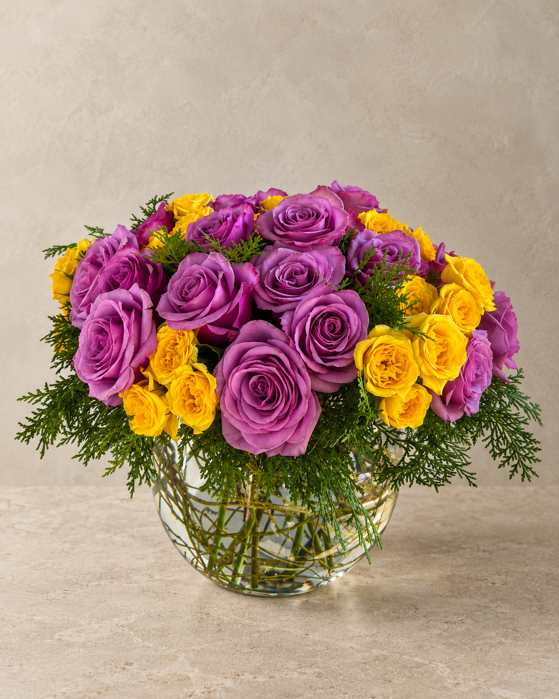 A vibrant flower bouquet featuring purple and yellow roses with green foliage, arranged in a clear glass vase on a light beige stone surface against a textured beige wall.