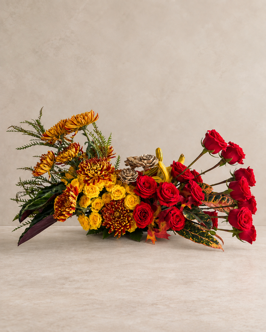 Full frontal view of a wide autumn arrangement with bronze chrysanthemums, yellow spray roses, dried rosettes, striped gourds and long-stemmed red roses.