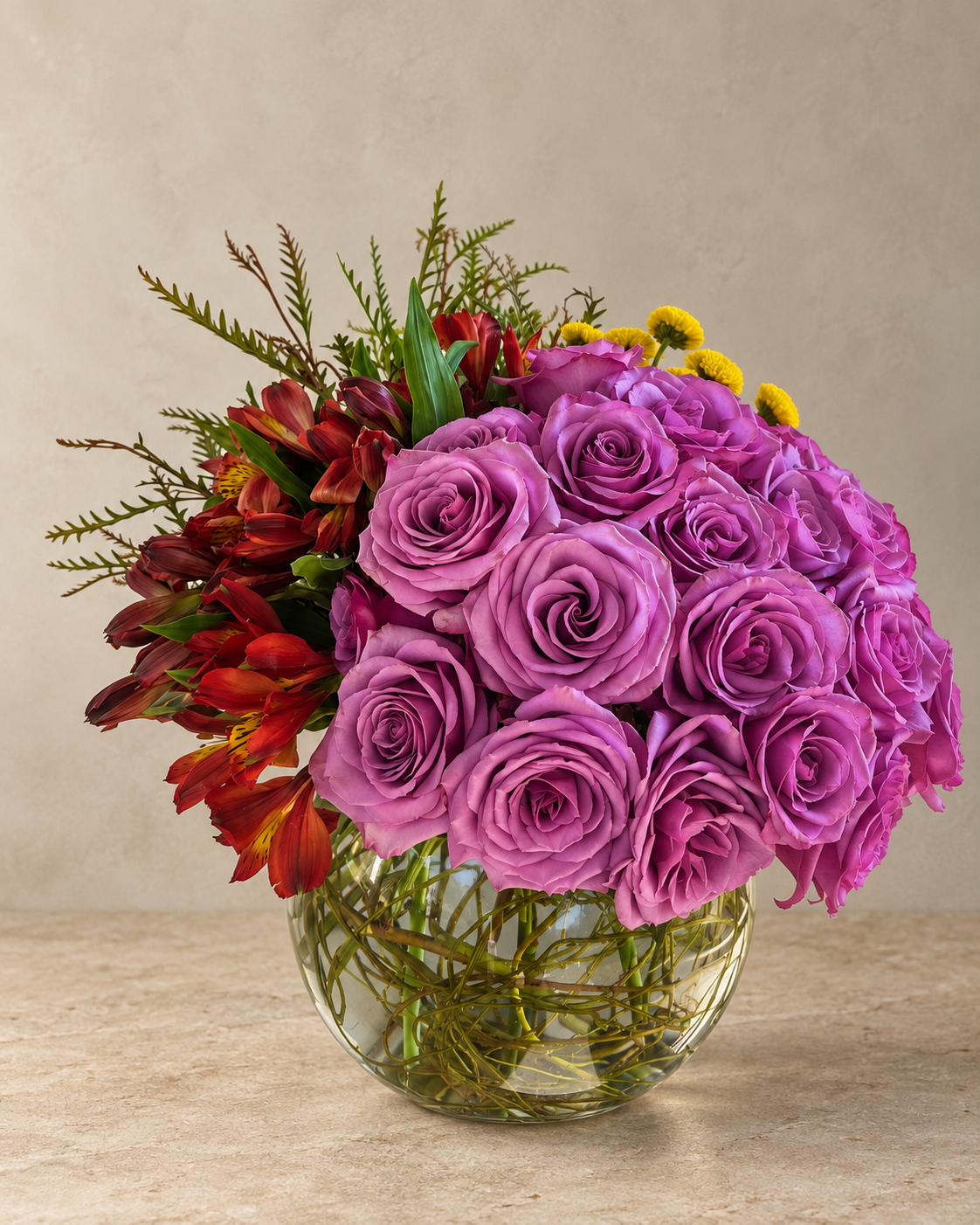 A vibrant flower bouquet featuring a large cluster of purple roses, red alstroemeria, and small yellow daisies arranged in a round clear glass vase on a beige surface against a neutral beige background.
