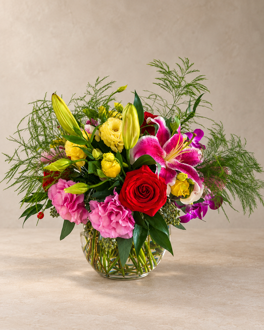 A vibrant mixed floral arrangement in a clear glass vase, featuring a red rose, pink carnations, yellow lilies, and pink lilies of the valley, set against a neutral beige background.