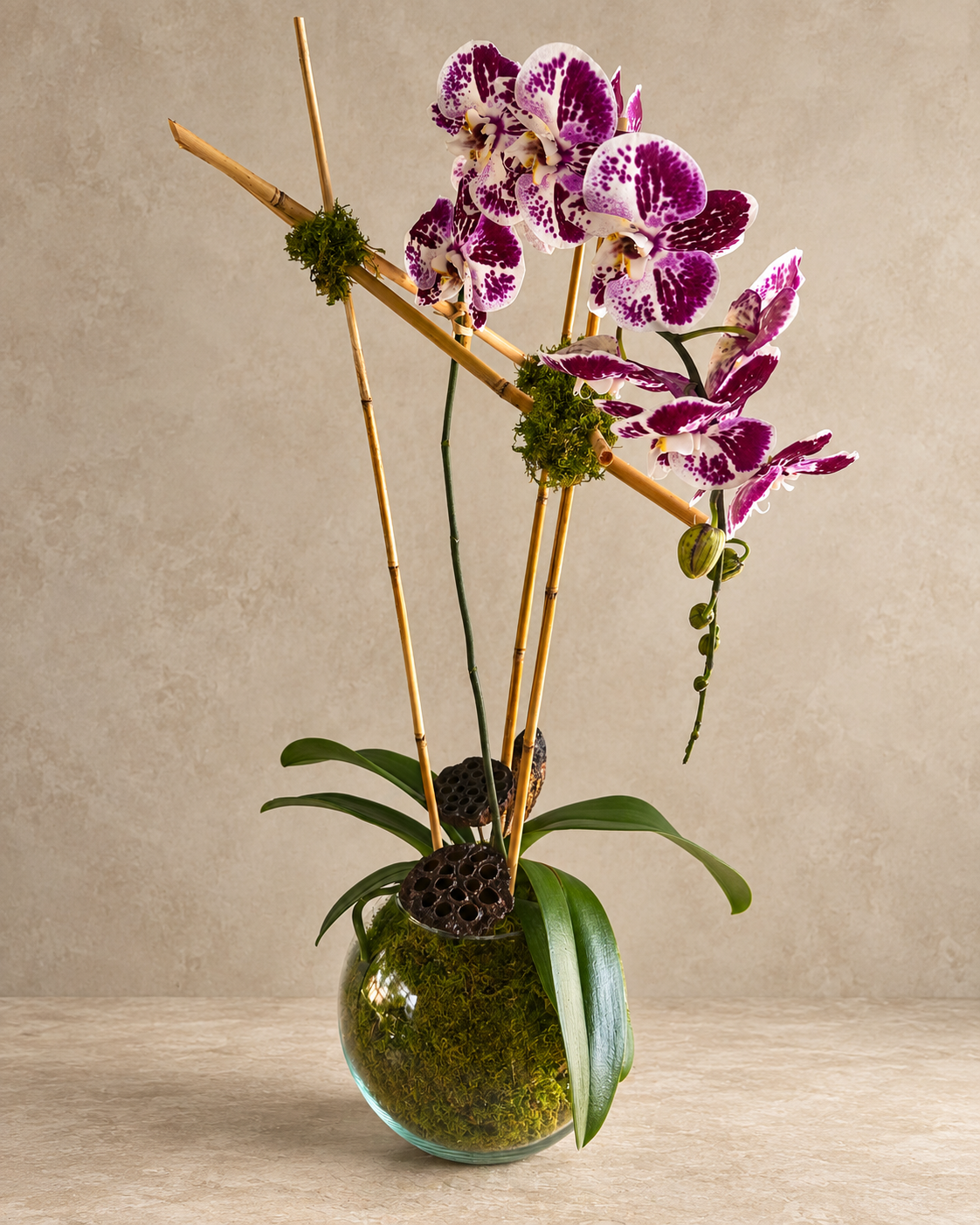 A decorative floral arrangement featuring purple and white spotted orchids with green leaves, mounted on natural bamboo stakes and mossy accents, set in a round glass vase filled with green moss, on a beige surface against a textured beige wall.