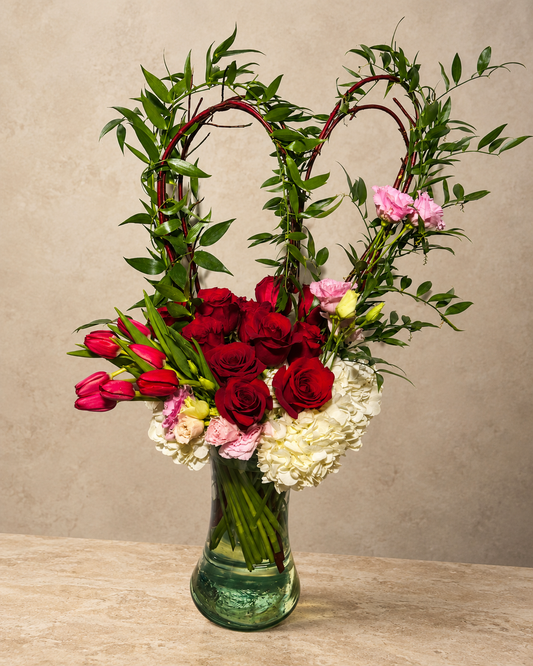Heart-shaped arrangement with red roses, red tulips, white hydrangeas and pink blooms in a green glass vase.