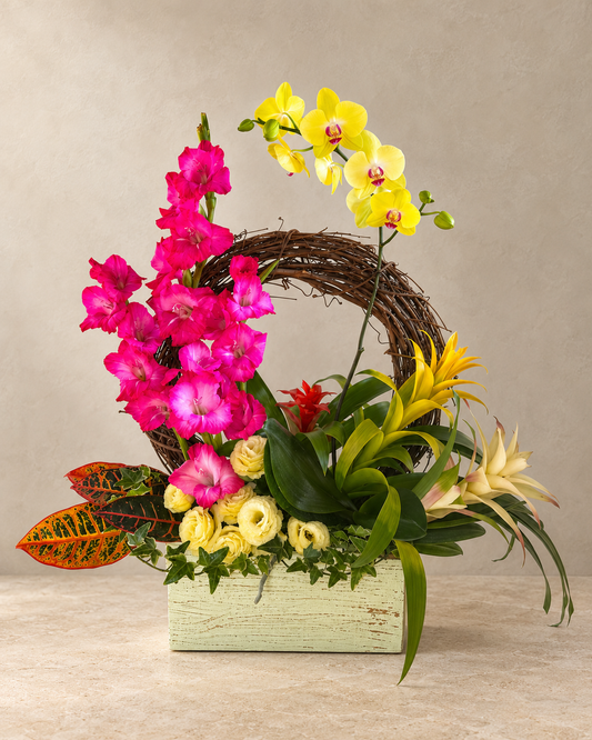 A vibrant floral arrangement in a light green rustic wooden box, featuring bright pink gladiolus, yellow orchids, yellow lilies, green foliage, and a brown grapevine wreath, set against a neutral beige background.