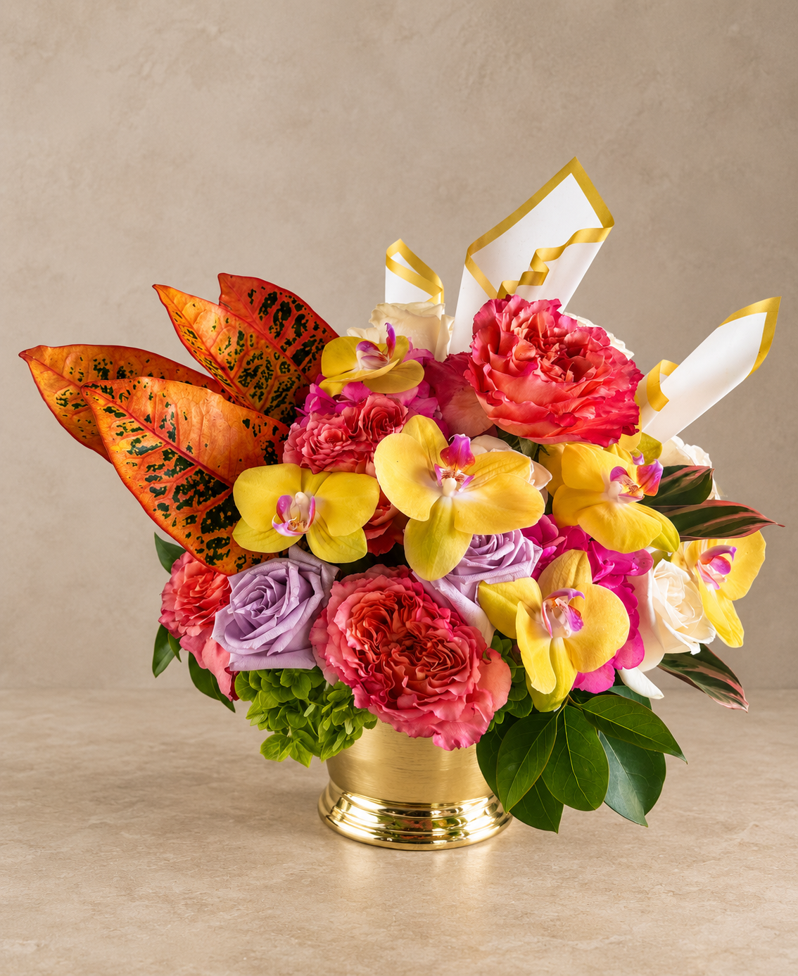 A vibrant floral arrangement in a gold pot, featuring yellow and pink orchids, pink and purple roses, and orange leaves with black speckles, set against a neutral beige background.