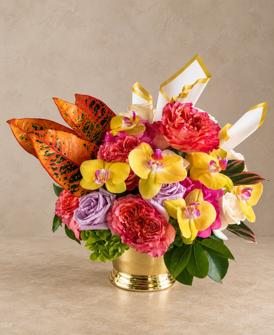 A vibrant floral arrangement in a gold pot, featuring yellow and pink orchids, pink and purple roses, and orange leaves with black speckles, set against a neutral beige background.
