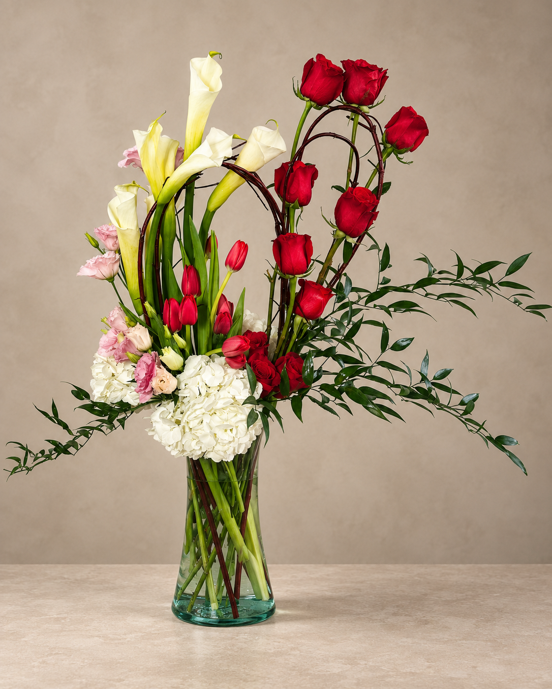 Full view of Souls in Love with sculptural red roses, white calla lilies, hydrangea and extended ruscus greenery in a clear cylinder vase.
