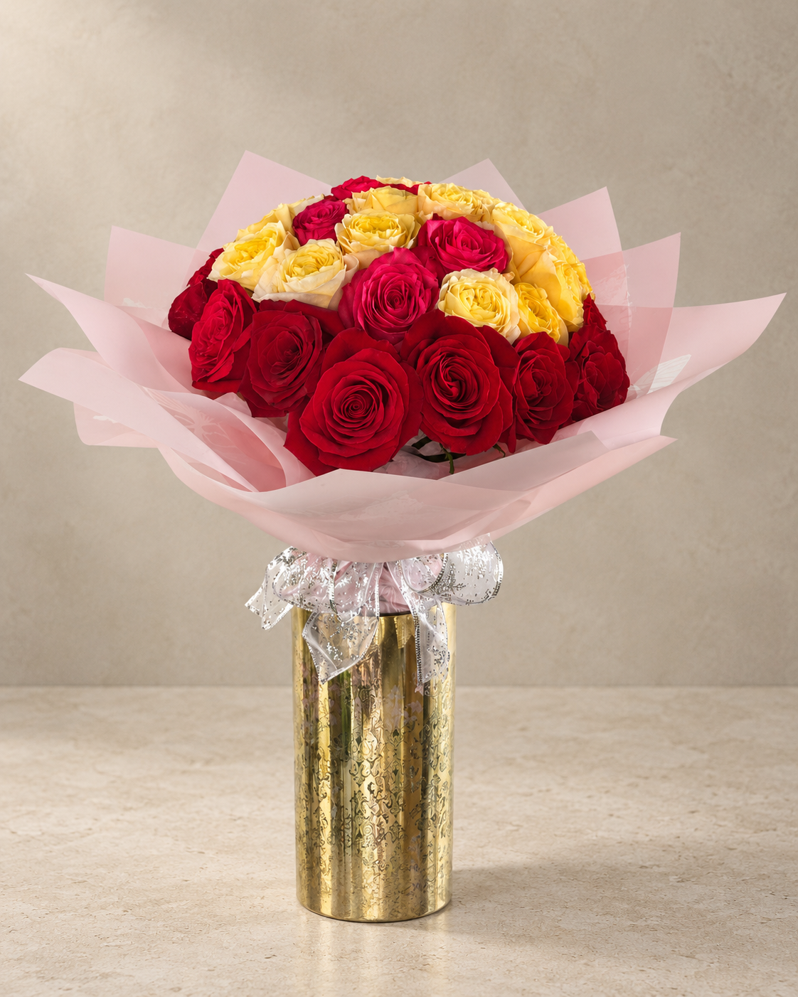 Full-length studio view of fresh deep-red and buttery-yellow roses wrapped in translucent pink paper, tied with a silver-edged bow and arranged in a tall gold vessel.