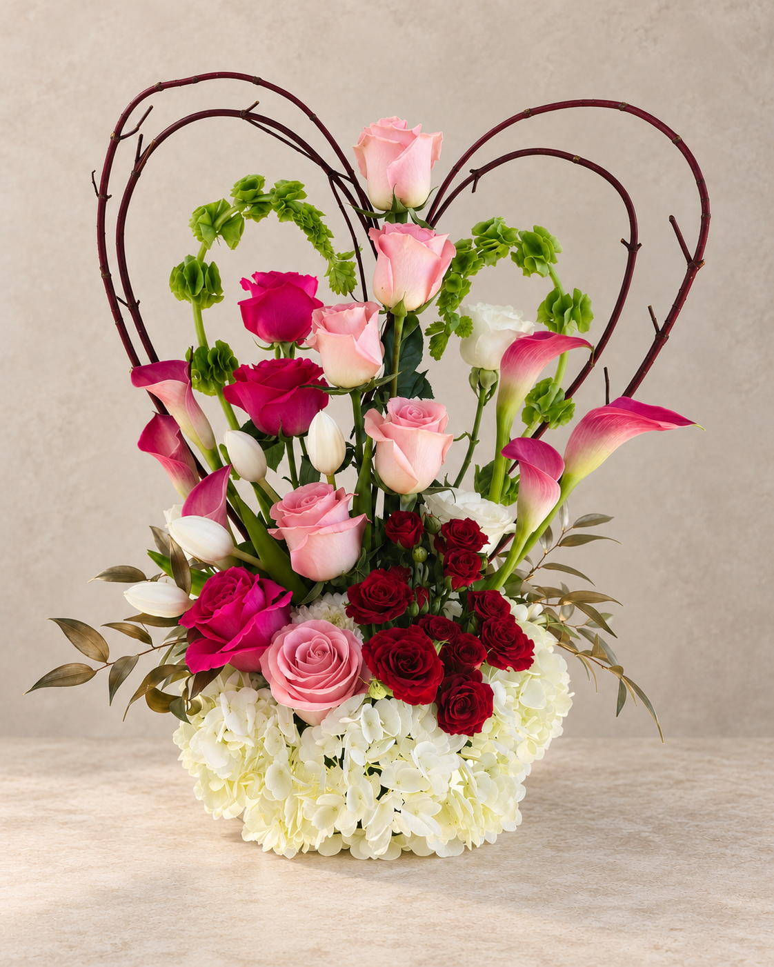 A heart-shaped floral arrangement featuring pink and red roses, white and pink calla lilies, white hydrangeas, and green buds, framed by dark brown vine arches on a neutral beige background.