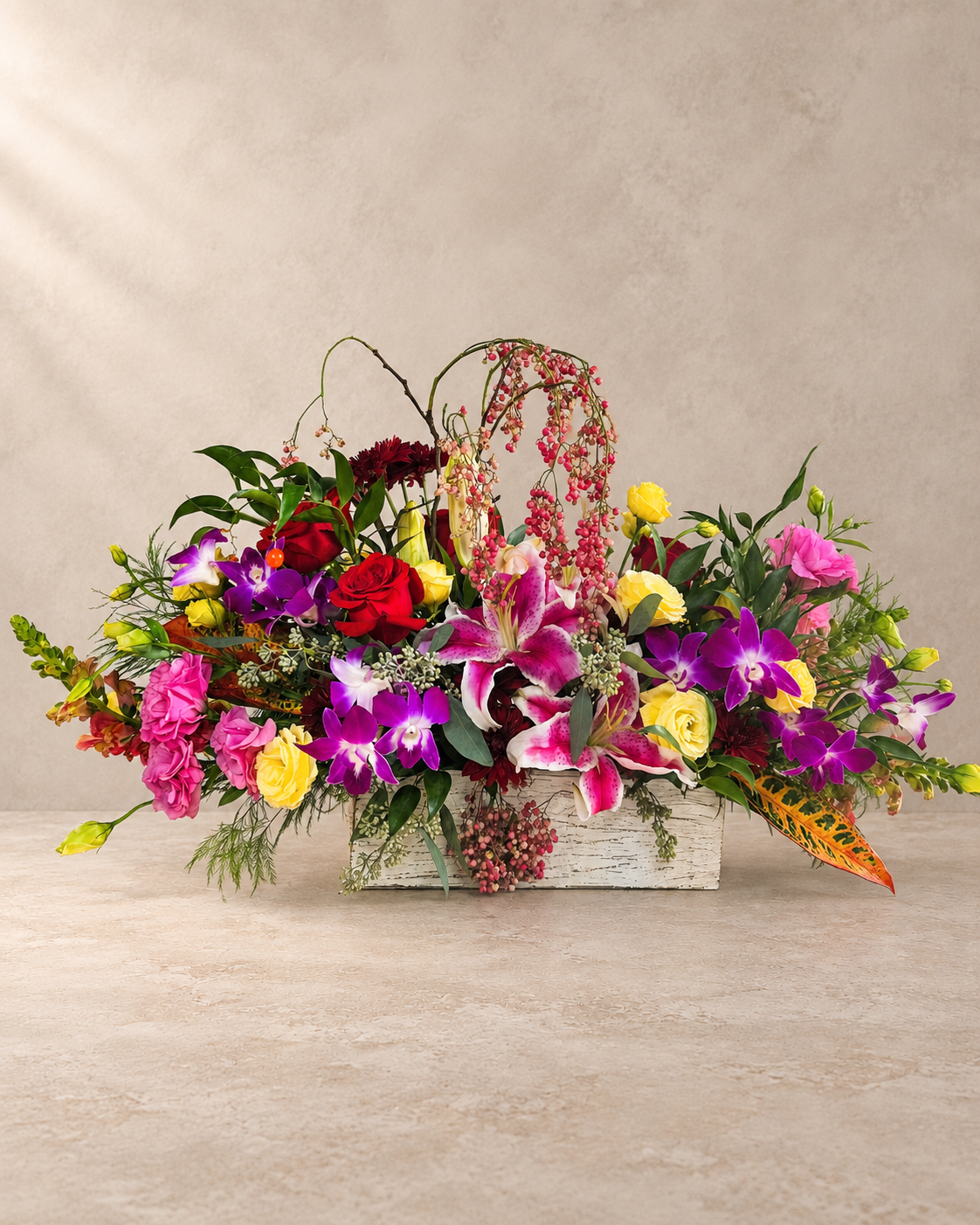 A vibrant floral arrangement in a rustic white wooden basket, featuring purple orchids, pink roses, yellow carnations, red roses, and green foliage, set against a neutral textured beige background.