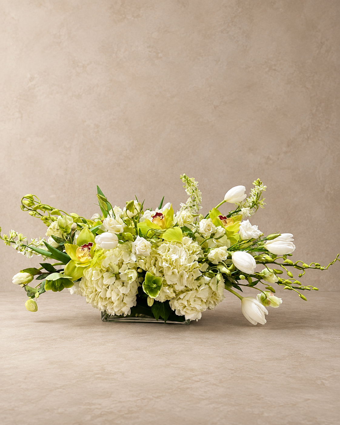 Chartreuse Green Delight low centerpiece with white hydrangea, chartreuse cymbidium orchids, white tulips and spray roses in a clear rectangular vase.