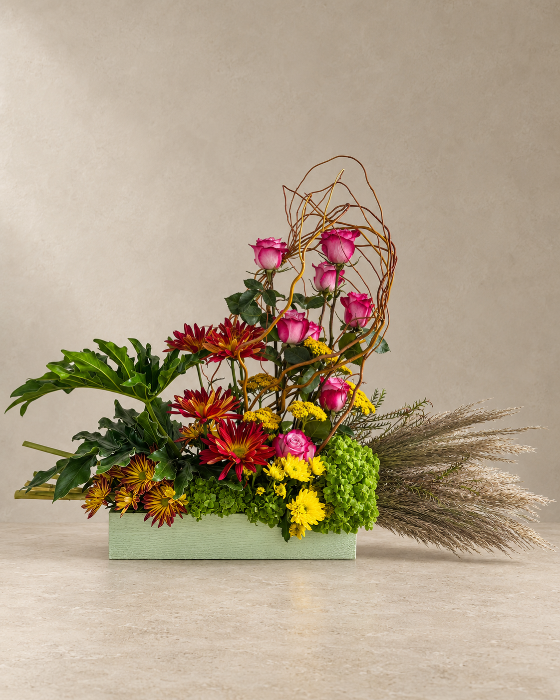 Wide asymmetrical arrangement with seven pink roses, red-bronze chrysanthemums, yellow flowers, green hydrangea, curly willow and sweeping ornamental grass in a sage rectangular vessel.