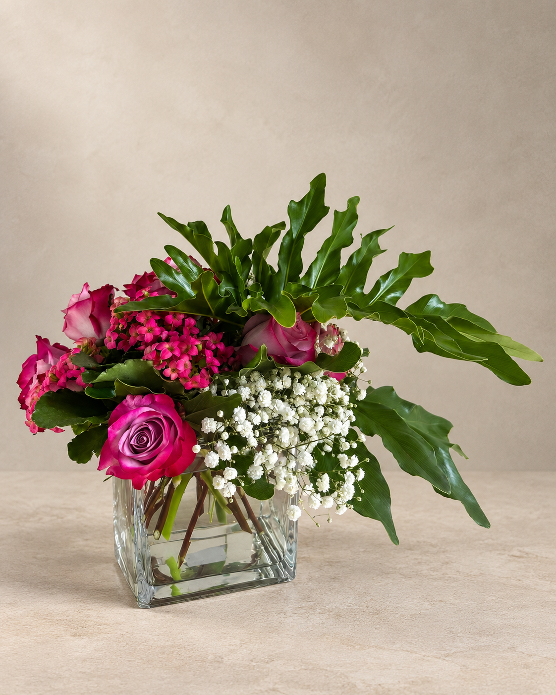 A floral arrangement in a clear square glass vase, featuring pink roses, bright pink blossoms, green leaves, and white baby's breath, set against a neutral beige background.