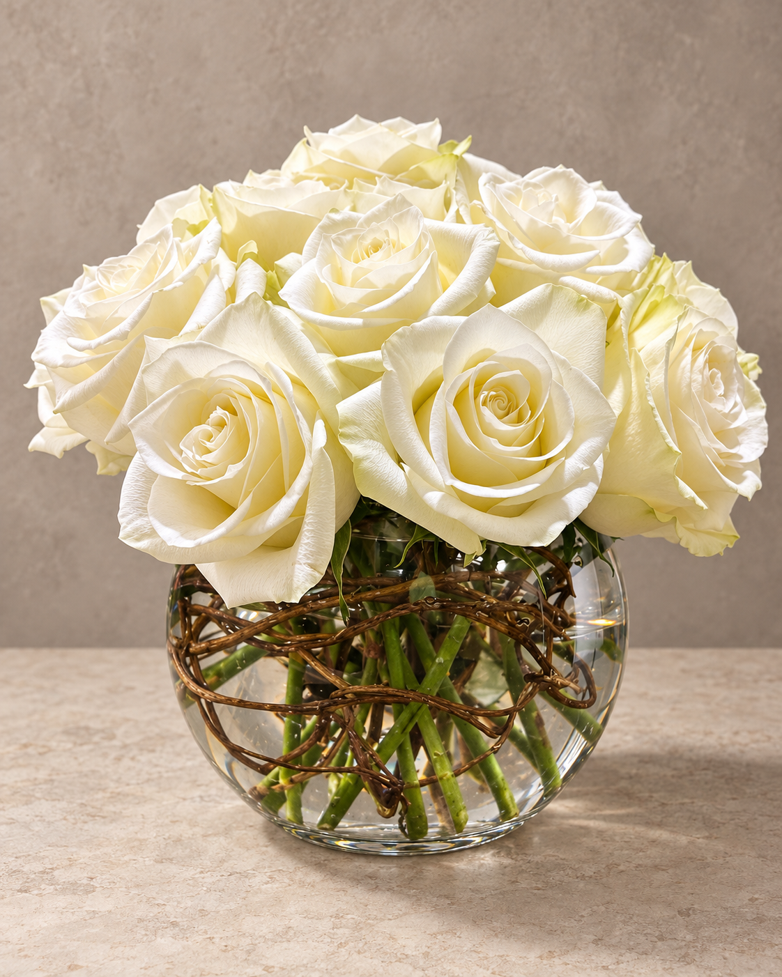 Full studio view of ivory-white roses arranged in a clear spherical glass vase with visible green stems and looping brown vine beneath the blooms.