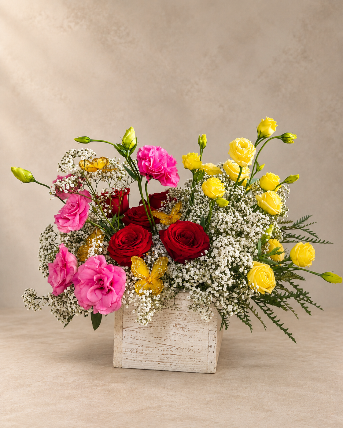 A vibrant floral arrangement featuring pink carnations, red roses, yellow lisianthus, and white baby's breath in a rustic white wooden box, set against a neutral beige background.
