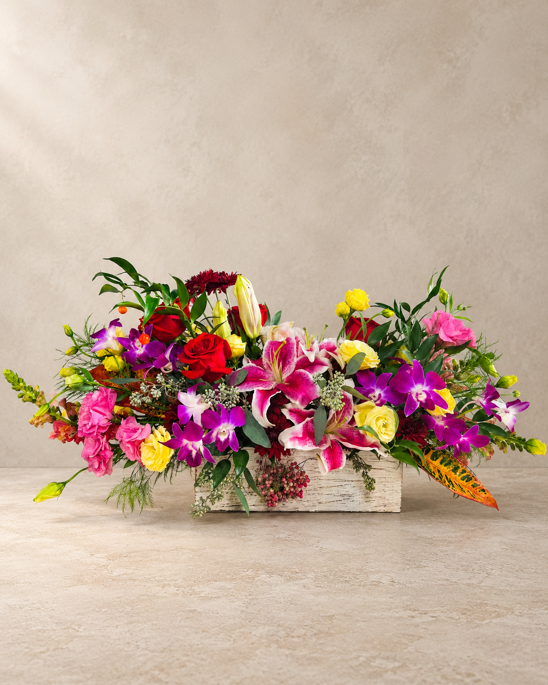 A vibrant floral arrangement featuring a mix of pink, red, yellow, and purple flowers with green foliage, presented in a rustic white-washed wooden box on a neutral beige background.