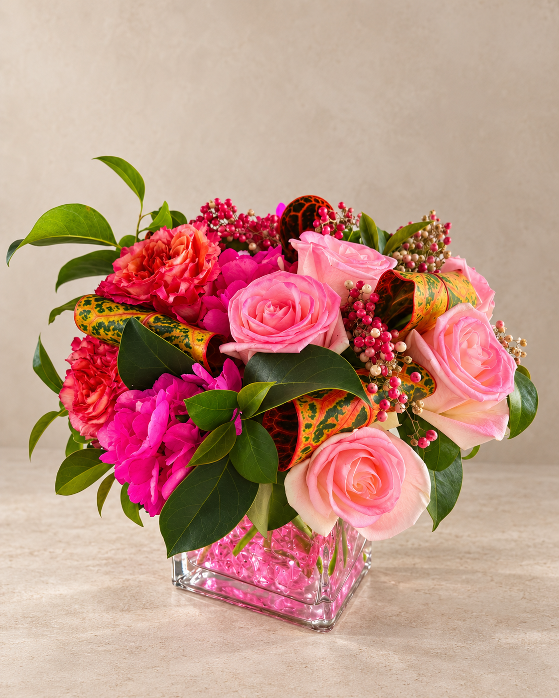 Centered studio view of a rounded pink floral arrangement with blush roses, magenta hydrangea, coral blooms, berry sprays and colorful croton foliage.
