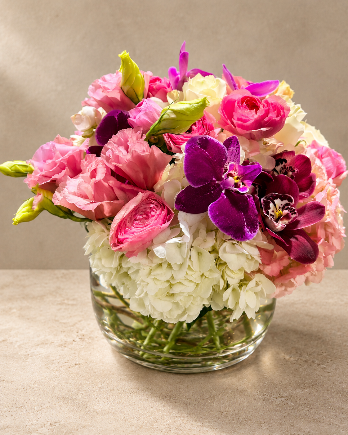A vibrant floral arrangement featuring pink and white flowers with deep purple orchids, presented in a clear glass vase on a neutral beige surface.