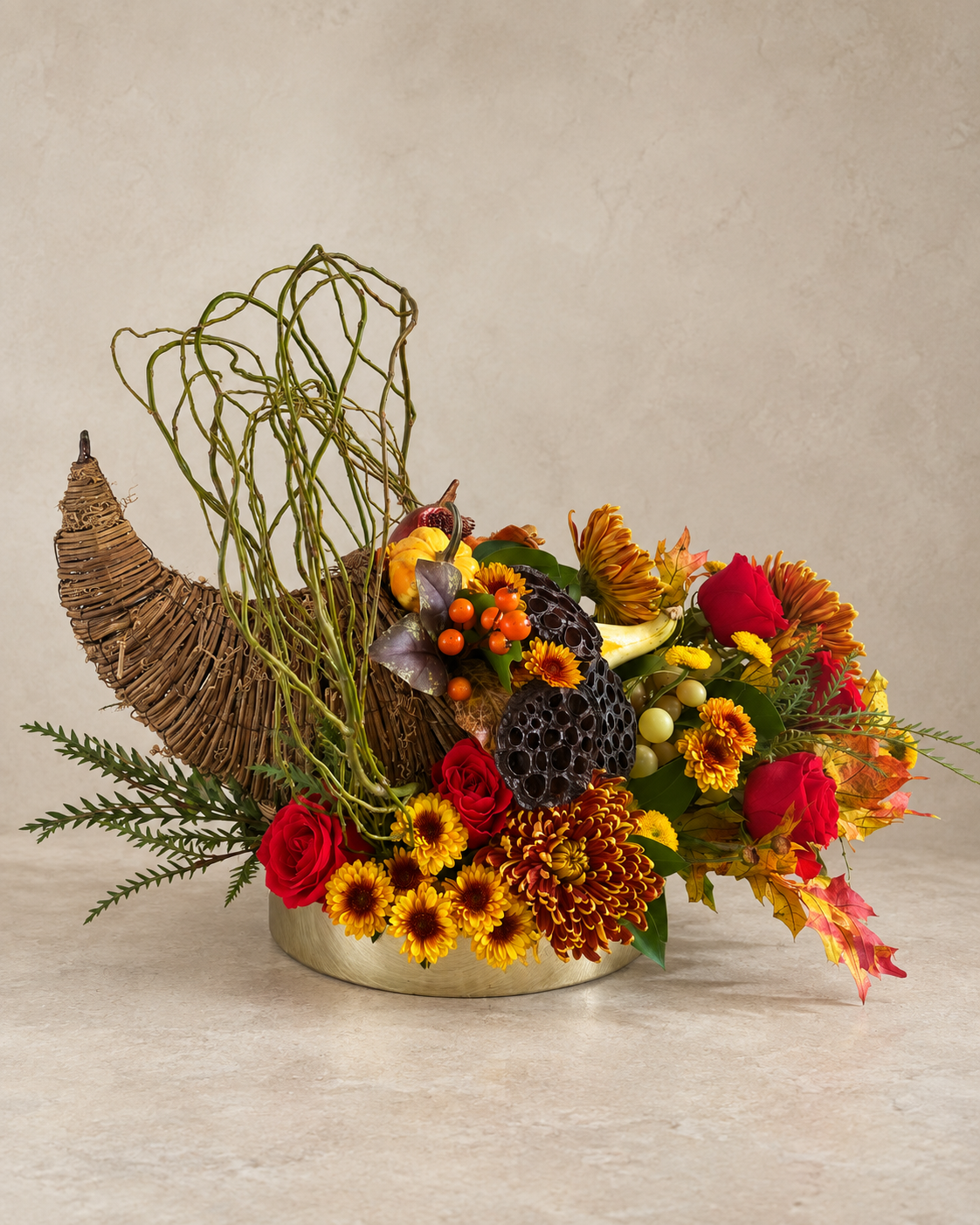 Elevated view of a cornucopia arrangement with red roses, bronze chrysanthemums, lotus pods, green grapes, gourds, orange berries and cascading autumn leaves.