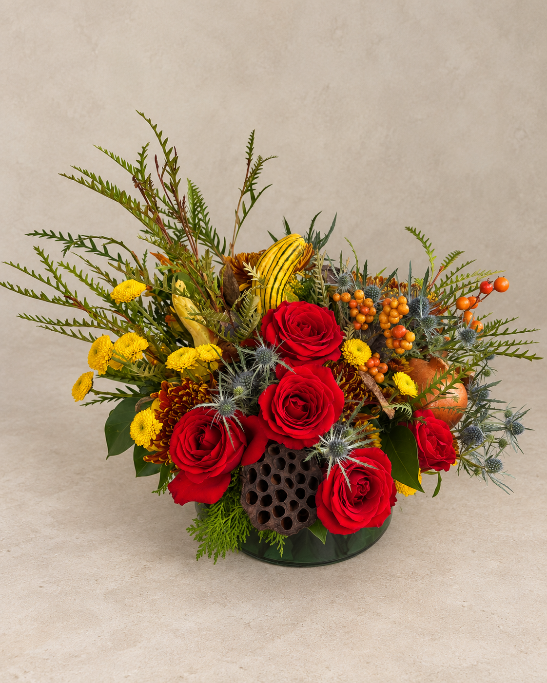 Elevated view of red roses, autumn chrysanthemums, yellow button flowers, thistle, berries, gourds and a dried lotus pod in a low green glass vase.
