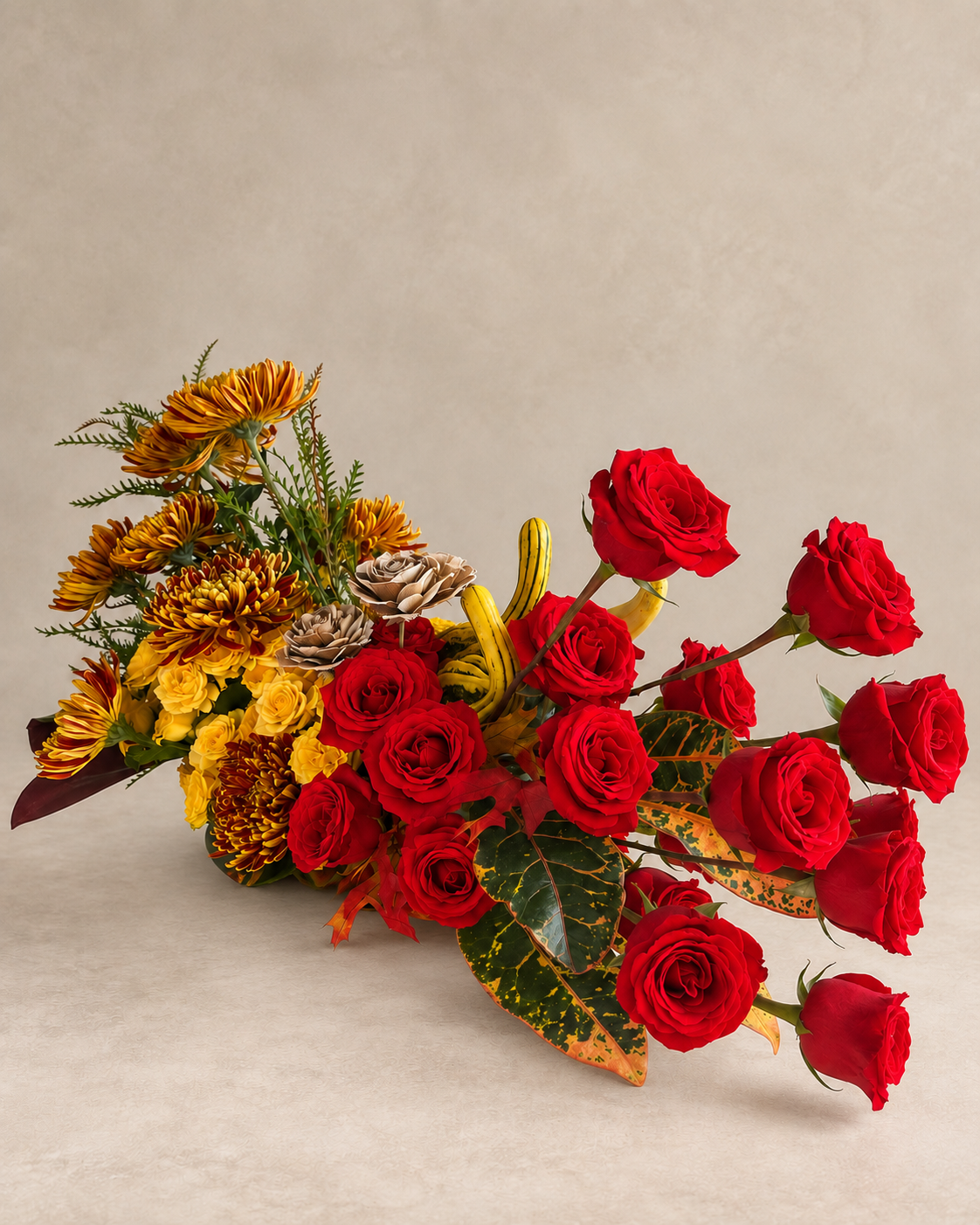 Elevated side view of red roses sweeping outward from bronze chrysanthemums, yellow spray roses, striped gourds, croton leaves and autumn foliage.