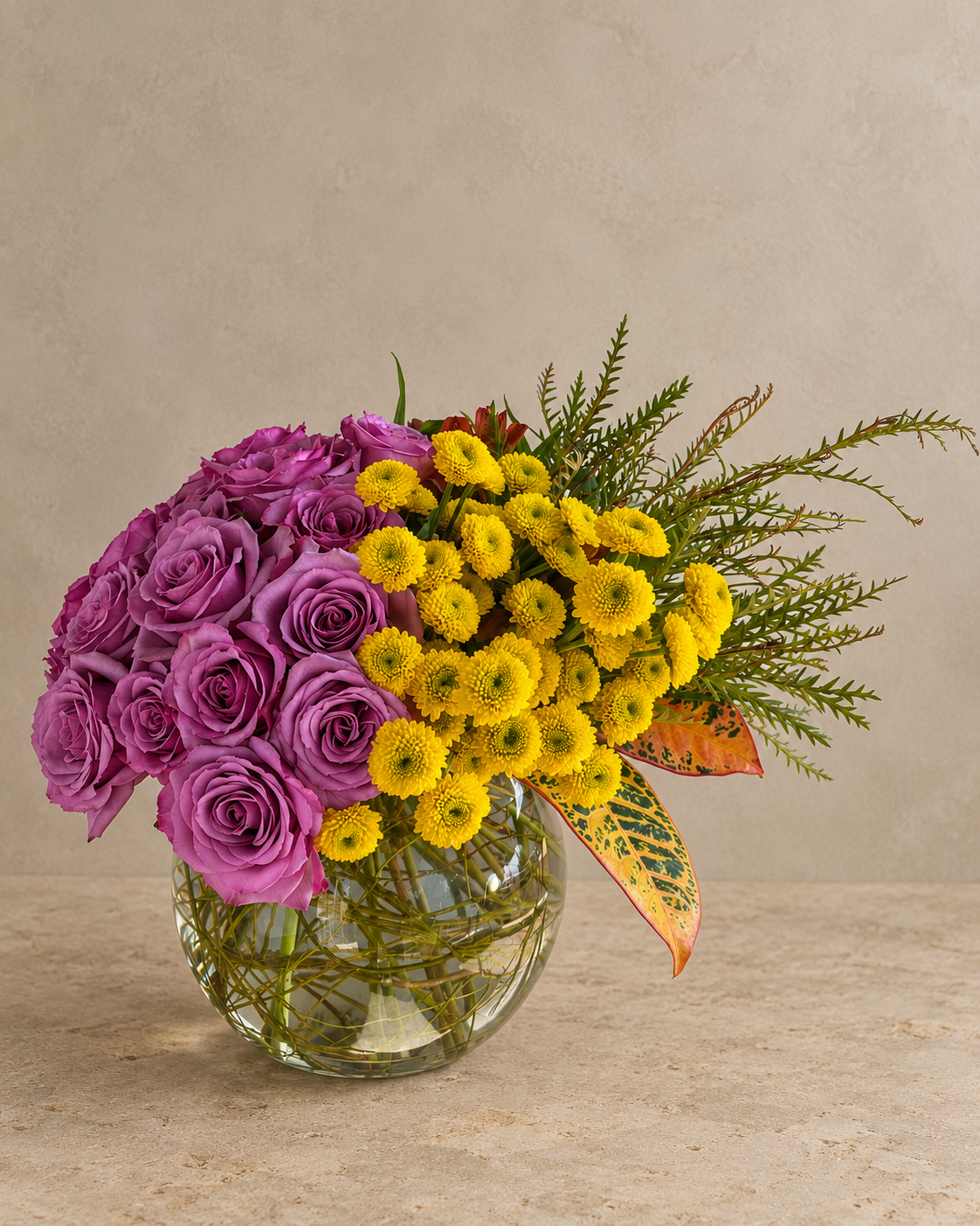 A floral arrangement featuring a cluster of purple roses and bright yellow daisy-like flowers in a clear round glass vase, with green foliage and a small orange accent leaf, set against a neutral beige textured background.