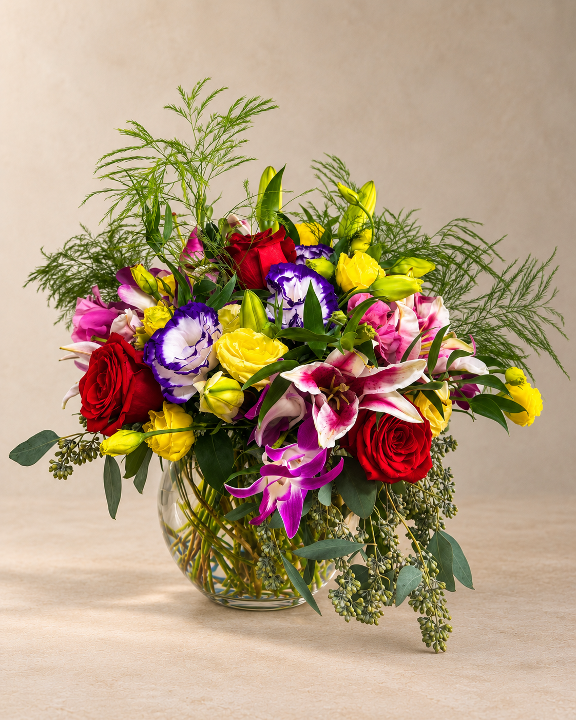 A vibrant mixed floral arrangement in a clear glass vase, featuring red and yellow roses, purple and white lilies, yellow tulips, and green foliage, set against a neutral beige background.