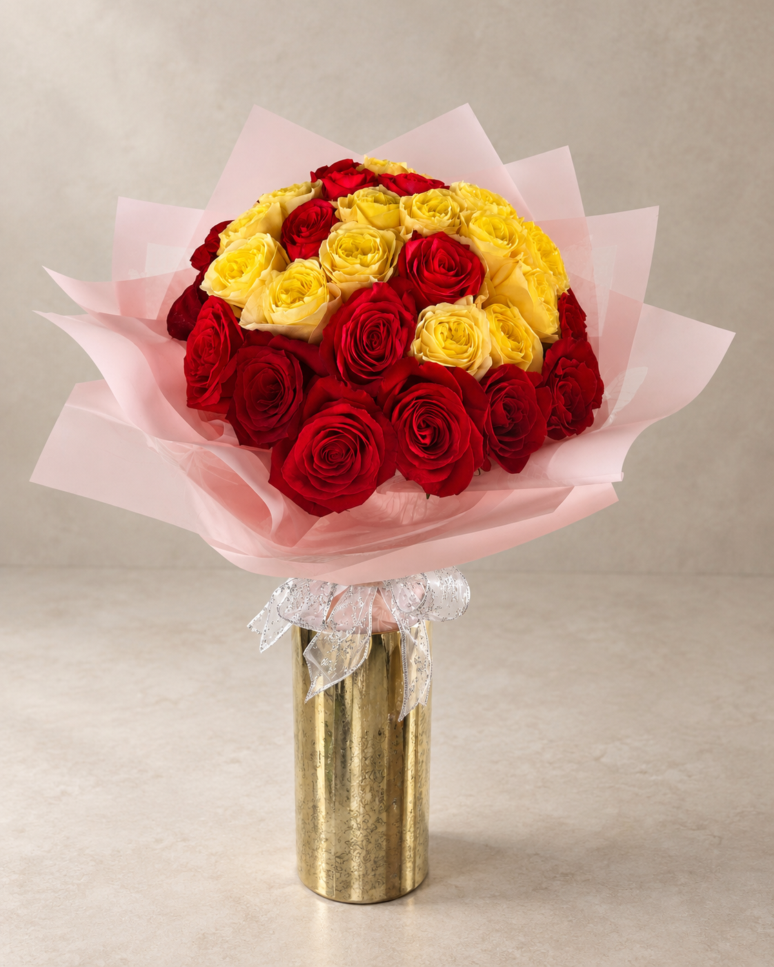 Elevated studio view of fresh red and yellow roses in layered pink wrapping, finished with a silver-edged bow and tall cylindrical gold vase.