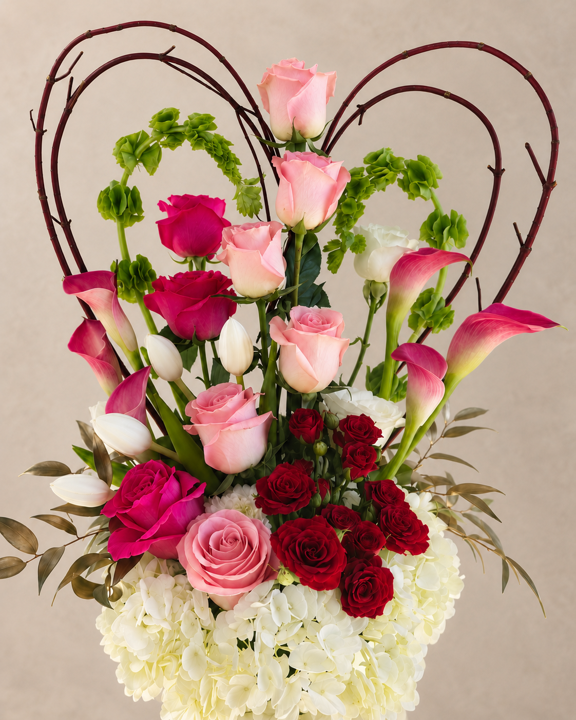 A heart-shaped floral arrangement featuring pink and red roses, pink and white calla lilies, green foliage, and a heart-shaped twig frame, set against a plain beige background.
