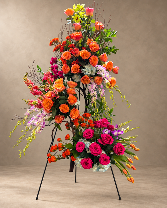 Manzanita Branch Clusters standing arrangement with orange roses, green and lavender orchids, hydrangea and hot-pink roses on a black easel.